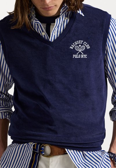 Sötétkék, strukturált mellény V nyakkivágással, fehér "Racquet Club Polo NYC" hímzéssel, egy kék és fehér csíkos ing felett viselve.