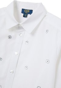 Camicia bianca con colletto classico, decorata con piccoli rivetti metallici argento a forma di fiore. Il tessuto ha una consistenza liscia.