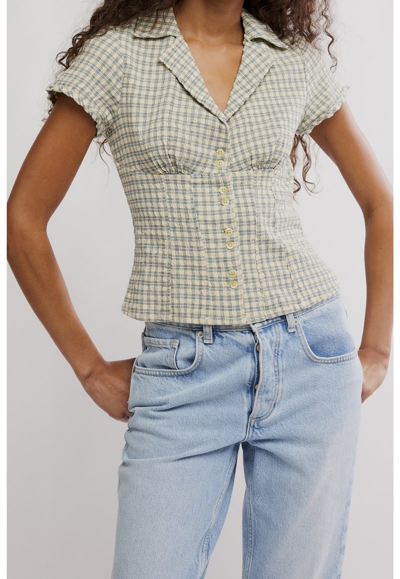 Femme portant un chemisier à manches courtes à carreaux jaunes avec boutons, associé à un jean taille haute bleu clair, mains reposant sur les hanches.