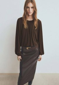 Blusa marrón de manga larga con escote fruncido, combinada con una falda lápiz de cuero marrón y un cinturón a juego con detalle metálico.