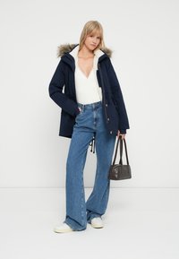 Parka blu navy con cappuccio foderato in pelliccia, top bianco a coste e jeans a vita alta azzurri a gamba larga. Una borsa nera e sneakers bianche completano l'outfit.