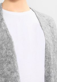 Vaaleanharmaa, teksturoitu neuletakki, joka on valmistettu pehmeästä materiaalista. Se on avonainen edestä ja pitkähiha, käytetty yksinkertaisen valkoisen T-paidan päällä.