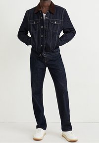 Man som bär en mörk denimjacka med brunt krage, matchande mörka denimjeans, vit inre skjorta och vita sneakers, står med händerna i fickorna.