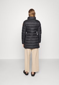 Sort dunjakke med høj krave og quiltet design, med en orange logo-patch. Modellen har beige bukser med vide ben og mørkt fodtøj på.