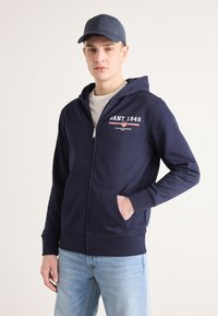 Sweat à zip bleu marine avec poches avant, poignets et ourlet côtelés. Présente le logo "GANT 1949" en blanc et rouge sur la poitrine. Porté avec un jean bleu clair.