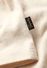 Beige fabric with visible stitching and a small black tag embroidered with "Speeday" in gold thread.