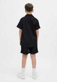 Camisa negra de manga corta y pantalones cortos a juego, hechos de tela texturizada, combinados con zapatillas blancas y calcetines blancos. Fondo liso.