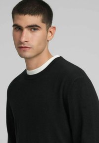 Zwarte gebreide trui met een ronde halslijn en een subtiel logo-accent. Zachte textuur, reguliere pasvorm, over een wit overhemd gedragen.