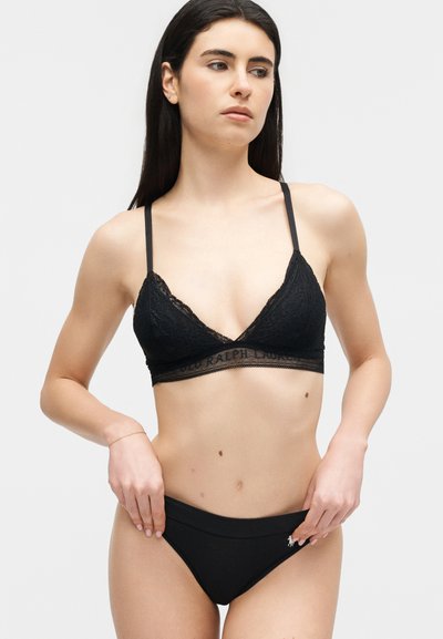 Γυναίκα με μακριά σκούρα μαλλιά, που φοράει μαύρο δαντελένιο μπραλέτο και μαύρο εσώρουχο σε στυλ μπικίνι, κοιτάζοντας ελαφρώς προς το πλάι.