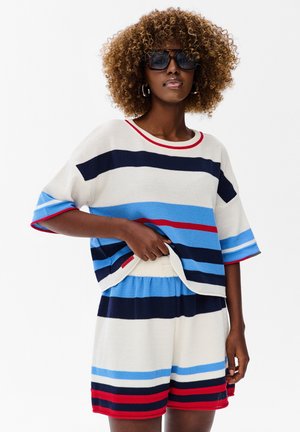 Kobieta z kręconymi włosami, nosząca ciemne okulary przeciwsłoneczne i dopasowany dzianinowy strój z poziomymi paskami w kolorach granatowym, niebieskim, białym i czerwonym.