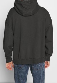 Felpa con cappuccio grigio scuro, dal taglio rilassato, con cuciture visibili e polsini a costine, abbinata a jeans blu con una texture di denim sbiadito.