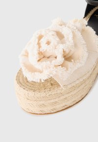 Sandalia de plataforma en crema con un acento de flor de tela texturizada, suela de yute trenzado y una suave correa negra en el tobillo.