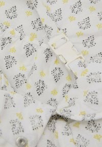 Vitt tyg med ett upprepat mönster av svarta och gula blommönster, med ett plastspänne synligt vid kragen.