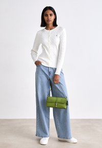 Witte kabelgebreide trui, lichtblauwe wijde jeans en een groene leren handtas met een gestructureerd ontwerp en zilveren hardware. Witte sneakers.