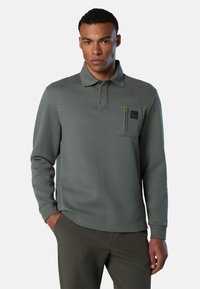 Polo in tessuto verde scuro con maniche lunghe, colletto e tasca con zip sul petto sinistro con una toppa logo.