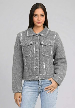 Femme portant une veste en laine grise boutonnée avec des motifs blancs brodés et deux poches poitrine, associée à un jean bleu clair, sur un fond gris.