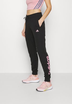 Femme portant un jogging Adidas noir avec un logo rose et des chaussures de sport roses, debout avec une main dans la poche sur un fond blanc.