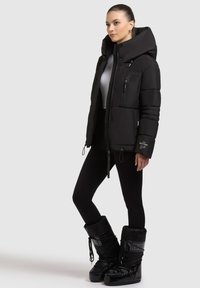 Sort foret jakke med høj krave, der har en lynlåsfront og flere lynlåslommer. Modellen bærer sorte leggings og glansfulde sorte støvler.