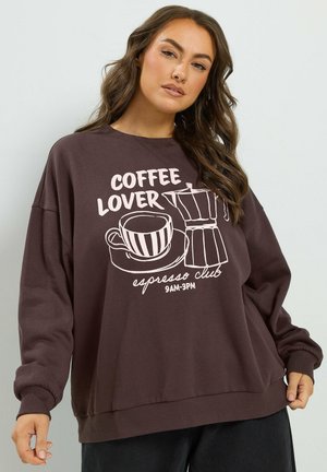 Femme portant un sweat-shirt marron avec le texte "Coffee Lover espresso club 9AM-3PM" et des illustrations d'une tasse à café et d'une cafetière.