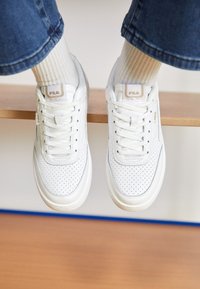 Sneaker bianchi in pelle con dettagli perforati, lacci piatti e un'etichetta con logo beige. Indossate con calzini bianchi a coste, posizionate su una superficie di legno.