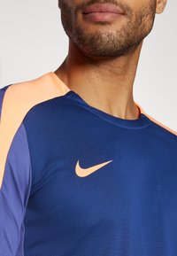 Mornarska športna majica s kratkimi rokavi, z breskevimi poudarki na ramenih in majhnim oranžnim logotipom Nike na prsih. Tekstura je gladka.