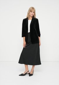 Melna samta žakete virs balta krekla, apvienota ar garu melnu kleitu ar baltiem punktiņiem un melnām slingback kurpēm ar loku akcentiem.