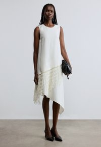 Rochie albă fără mâneci, cu tiv asimetric, având detalii cu franjuri în partea de jos. Modelul ține o geantă de mână din țesătură neagră.
