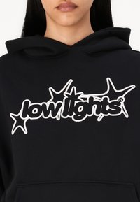 Μαύρο φούτερ με λευκό γραφικό λογότυπο που γράφει "low lights" με αστεράκια, φτιαγμένο από μαλακό ύφασμα. Φαίνεται unisex με άνετη εφαρμογή.