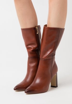 Bottes en cuir marron montant jusqu'au milieu du mollet, avec des bouts pointus, des fermetures éclair latérales et des talons carrés, portées sur des jambes nues sur un fond blanc.