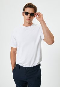 Camisa de algodón blanca de manga corta, combinada con pantalones azul marino. Modelo usando gafas de sol redondas y oscuras, sujetándose las sienes con la mano derecha.