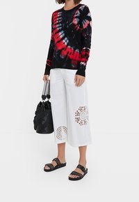 Pull tie-dye noir et rouge à manches longues, assorti d'un pantalon blanc à jambes larges avec des accents en dentelle, ainsi que des sandales noires et un sac.