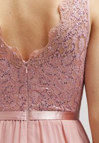 Robe rose pâle avec un dos en dentelle agrémenté de sequins, un décolleté en V profond et une ceinture en satin avec des plis. Fermeture éclair visible.