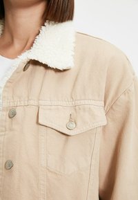 Béžová džínsová bunda s huňatým bílým límcem, se stříbrnými knoflíky a kapsou na prsou. Strukturovaná tkanina s jemným povrchem.