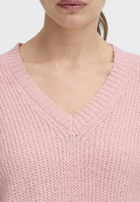 Maglione lavorato a maglia di un leggero rosa, con scollatura a V. Il tessuto strutturato mostra un motivo di maglia allentata e orlo a coste. Materiale morbido e leggero.
