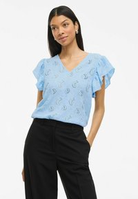 Gaiši zila blūze ar V veida kakla izgriezumu un īsām volānu piedurknēm, ar teksturētu rakstu. Apvienota ar melniem pielāgotiem biksēm.