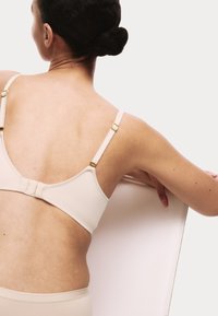 Beige beha met verstelbare bandjes en gouden details, met een gladde textuur en zachte randen. Model trekt aan een stuk stof achter zich.