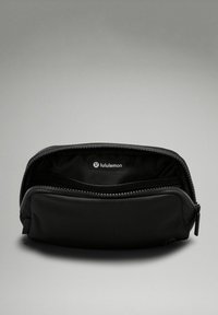 Pochette zippée noire avec un extérieur lisse et des côtés texturés. L'intérieur comporte une étiquette de logo et un compartiment zippé séparé pour l'organisation.