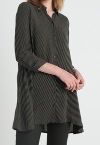 Blusa nera con bottoni, a pois bianchi, maniche a tre quarti, orlo curvo, tessuto morbido e vestibilità ampia.