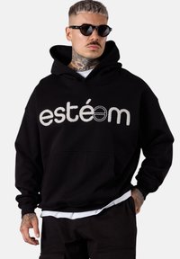 Hoodie noir avec poche kangourou à l'avant, comportant un logo brodé blanc "estéom". Le tissu semble doux avec une coupe décontractée et une capuche réglable.