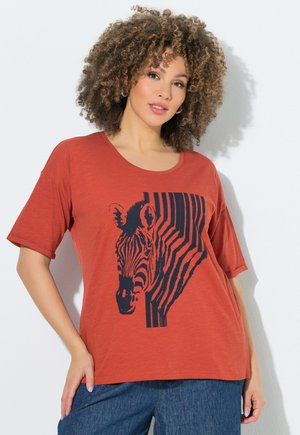 Kobieta z kręconymi włosami, ubrana w koszulkę w kolorze rdzy z granatowym nadrukiem zebry, połączoną z niebieskimi jeansami.