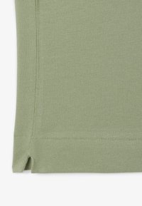 Tessuto lavorato a maglia verde chiaro con una superficie testurizzata, caratterizzato da spacchi laterali sull'orlo per maggiore flessibilità e una finitura pulita lungo i bordi.