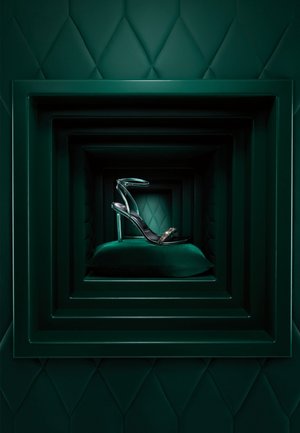 Sandale à talon noir avec des brides transparentes et des accents dorés, présentée sur un coussin en velours vert dans un cadre vert en couches.