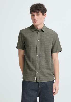 Młody mężczyzna z krótkimi ciemnymi włosami, ubrany w oliwkowo-zieloną teksturowaną koszulę na krótki rękaw z guzikami i ciemnoniebieskie dżinsy, stojący na jasnym tle.