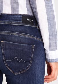 Vaqueros de mezclilla azul oscuro con un bolsillo trasero que presenta un diseño de costura curvada, parche de cuero de marca y detalle de costura en contraste.
