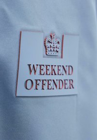 Tela blanca con un parche rectangular blanco que presenta un símbolo de corona y el texto rojo "WEEKEND OFFENDER" en relieve.