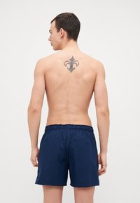 Mørkeblå svømshorts med elastisk talje, laget av lett stoff; har et dekorativt tatoveringsmotiv i form av en fleur-de-lis på baksiden.