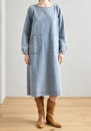 Person wearing a light blue denim midi dress with long sleeves and two front pockets, paired with tan leather mid-calf boots.