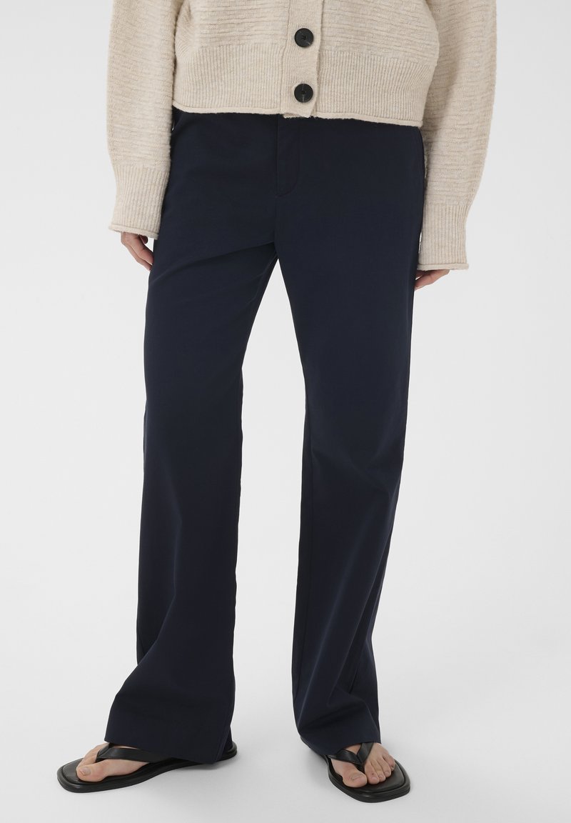 Personne portant un pantalon large bleu marine, un pull en maille beige boutonné et des tongs noires à bout carré, debout devant un fond uni.