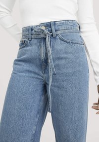 Vaaleansiniset farkut, joissa on korkea vyötärö, solkitys ja etutaskut. Kangas on hieman teksturoitu ja siinä näkyy ompeleet.