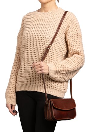 Borsa a tracolla in pelle marrone con chiusura a pattina, cuciture a contrasto e una lunga tracolla, tenuta da una persona che indossa un maglione beige con texture.